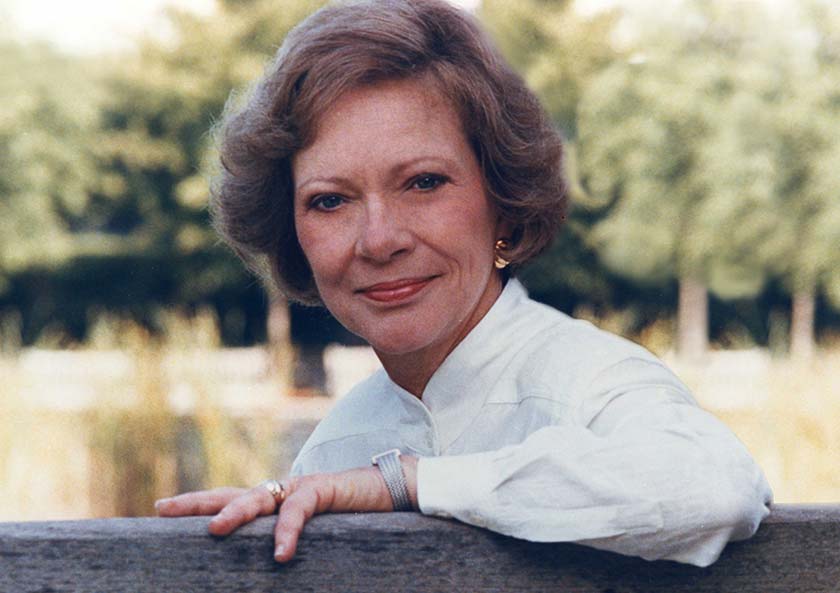 A woman leaning on a wooden fence in front of trees.