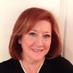 A smiling woman with red hair wearing a black top and pearl necklace.