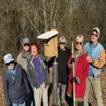 Six people outdoors holding birdhouses and tools.
